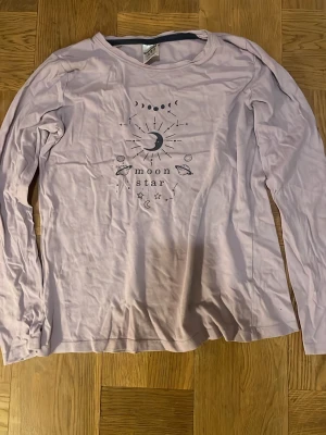 Ljuslila långärmad pyjamas från Kappahl - Ljuslila långärmad pyjamas topp från Kappahl med tryck på framsidan som visar måne, stjärnor och planeter samt texten 'moon star'. Rund halsringning och mjukt bomullsmaterial. Perfekt för dig som gillar astrologi och coola prints. Väl använd men i bra skick. Pris kan diskuteras.