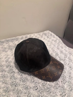 Louis Vuitton svart och brun keps - Säljer en snygg keps från Louis Vuitton med klassiskt monogrammönster. Kepsen är svart med brunt skinn på skärmen, båda delarna har LV-loggor och blommor. Justerbar baktill och riktigt lyxig vibe. Perfekt för dig som vill sticka ut med designeraccessoar.