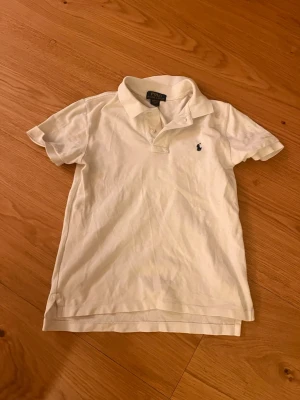 Vit pikétröja från Polo Ralph Lauren - GE PRIS FÖRSLAG!! Klassisk vit pikétröja från Polo Ralph Lauren med broderad logga på bröstet. Tröjan har korta ärmar, krage och knappar framtill. Snygg och enkel stil som passar till jeans eller shorts.