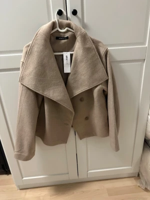 Beige kappa från Gina Tricot S helt ny med prislapp  - Snygg beige kappa från Gina Tricot i storlek S med bred krage och dubbelknäppning framtill. Jackan har en croppad och loose passform med långa ärmar och är tillverkad i mjukt material som ger en lyxig känsla. Perfekt för dig som gillar stilrena och trendiga plagg. Säljer billigare ny pris 700kr 
