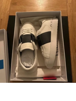 Valentino Open sneakers vit/svart - Säljer ett par Valentino Open sneakers i vitt skinn med bred svart rem över mitten. Snygga detaljer med nitar på hälen och platt sula med rutmönster undertill. Klassisk rund tå och snörning framtill. Perfekt för dig som gillar stilrena och lyxiga sneakers.