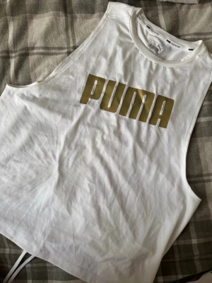 Vit linnetopp från Puma med snörning - Säljer en vit linnetopp från Puma med stort guldigt logotryck framtill. Toppen är ärmlös och har snygg snörning i ryggen som ger en cool detalj. Perfekt för träning eller chill dagar. Materialet är mjukt och lätt polyester.
