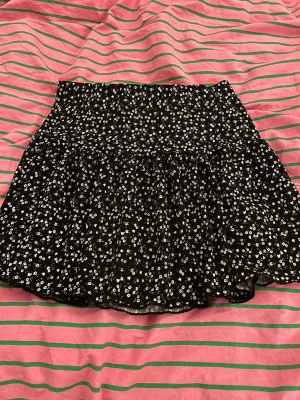  blommig kortkjol med volang - Säljer en supersöt svart kortkjol med små vita blommor och volangkant. Kjolen har en bred, smockad midja som ger en bekväm och snygg passform, streching så passar xs/s. Perfekt för sommaren🌻