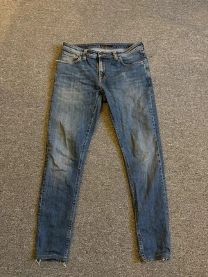 Blå skinny jeans från Nudie Jeans - Säljer ett par klassiska blå skinny jeans från Nudie Jeans i modellen W31 L32. Jeansen har en smal passform, fem fickor och snygga slitningar på benen. Tillverkade i mjukt jeansmaterial med orangea sömmar och Nudie-logga bak.