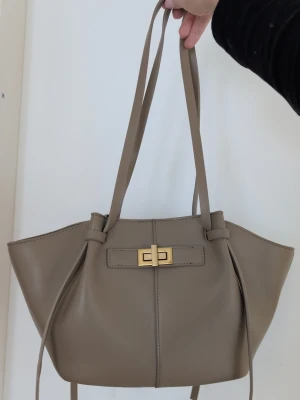 Beige axelväska med guldfärgat spänne - Snygg beige axelväska i skinnimitation med långa axelband och ett stilrent guldfärgat vridlås framtill. Väskan har en modern, strukturerad form med dekorativa band på sidorna och minimalistisk design. Perfekt för dig som gillar clean och trendig look. Använd ca 3ggr så typ nyskick