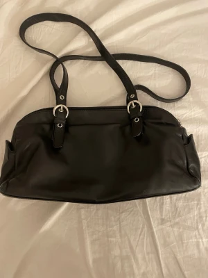 Svart axelväska från The Trend - Säljer en stilren svart axelväska från The Trend, tillverkad i Italien. Väskan är gjord i skinn och har flera fack med dragkedja, silverfärgade detaljer och justerbara axelremmar. Perfekt storlek för att få plats med allt du behöver och passar till många olika stilar.