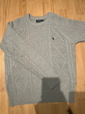 Grå stickad tröja från Polo Ralph Lauren - Snygg grå stickad tröja från Polo Ralph Lauren med klassiskt kabelstickat mönster och broderad logga på bröstet. Rund hals och ribbade muddar vid ärmslut och nederkant. Perfekt för dig som gillar stilrena och tidlösa plagg.