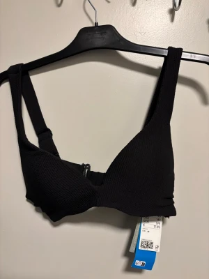 Svart ribbad bikiniöverdel - Svart bikiniöverdel med ribbad struktur och justerbara axelband. Modellen har en klassisk form med breda band och spänne bak. Perfekt för dig som gillar stilrena badkläder med lite extra stöd.