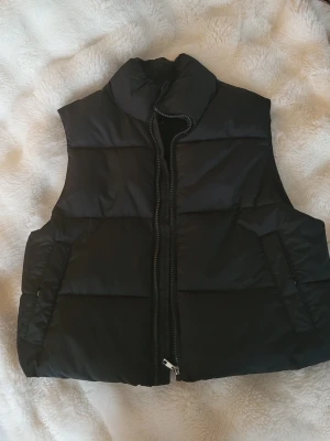 Svart dunväst  - Svart dunväst från 157 i storlek S. Västen har en hög krage och grov dragkedja framtill. Quiltad design med breda paneler och två sidofickor. Perfekt för lager-på-lager under kyliga dagar. Materialet är polyester och västen har en stilren, enkel look.