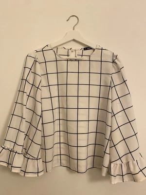 Rutig blus med volangärm - stl M - Säljer en vit blus med svart rutmönster och fina volangdetaljer på ärmarna. Blusen är använd men i bra skick. Lätt och luftig modell som passar perfekt både till vardags och mer uppklätt.