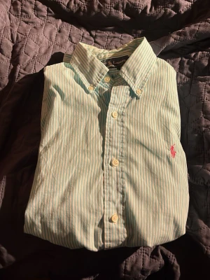 Randig skjorta från Ralph Lauren - Fräsch ljusblå och vit randig skjorta från Ralph Lauren med klassisk krage och knappar framtill. Skjortan har lång ärm och den ikoniska rosa logon broderad på bröstet. Perfekt för dig som gillar stilrena och snygga detaljer.