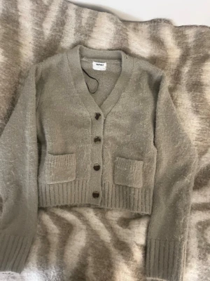 Beige stickad kofta från Monki - Mysig beige stickad kofta från Monki i storlek S. Koftan har v-ringning, fyra mörka knappar framtill och två praktiska fickor. Ribbstickade muddar vid ärmslut och nederkant ger en snygg touch. Perfekt för lager-på-lager under kyliga dagar.Den är grön typ