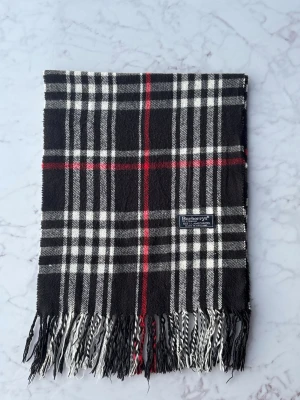 Rutig svartvit ullväst från Burberrys - Snygg väst från Burberrys i klassiskt rutigt mönster med svarta, vita och röda toner. Tillverkad i 100% lammull, mjuk och varm med fransar nedtill. Perfekt för dig som gillar tidlös design och vill sticka ut med ett ikoniskt plagg.