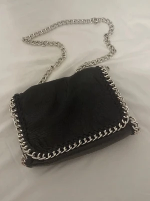 Svart axelväska med kedjedetaljer - Snygg svart axelväska med grov silverfärgad kedja som axelrem och runt kanterna. Väskan har en mjuk struktur och en cool, modern look som passar till många outfits. Perfekt för dig som gillar stilrena accessoarer med en twist.