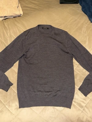 Mörkgrå stickad tröja från J.Lindeberg - Snygg mörkgrå stickad tröja från J.Lindeberg med rund hals och långa ärmar. Tröjan har en klassisk, normal passform och diskret logga på bröstet. 100% merinoull