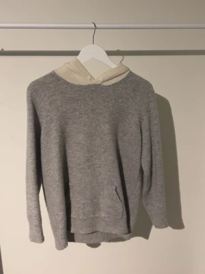 Grå hoodie merinoull  - Mysig grå hoodie med beige huva och långa ärmar. Tröjan har en klassisk känguruficka framtill och ribbade muddar vid ärmslut och nederkant. Perfekt för kyliga dagar och enkel att matcha med jeans eller mjukisbyxor.