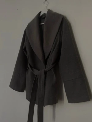 Grå kappa från BikBok med bälte - En grå/ljus grå kappa från bik bok jättefin, använd sparsamt. Tvättade den och fick någon fläck men vet ej om det går att få bort men de är inte speciellt märkbart när man har den på sig!