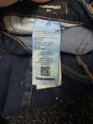 Dsquared2 Slim Jeans med färgstänk - Mörkblå slim jeans från Dsquared2 med coola slitningar och färgstänk på både fram- och baksida. Snygga kontrastsömmar och ett stort rött Dsquared2-märke bak. Tillverkade i Italien, klassisk femficksmodell och skinny passform.