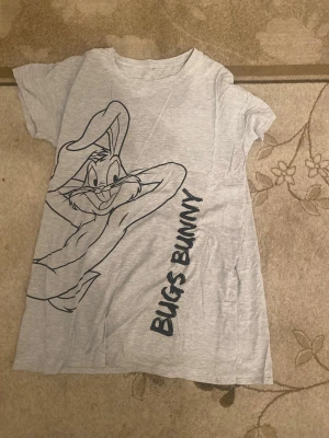 Grå Bugs Bunny klänning i bomull - Säljer en grå kortklänning med stor Bugs Bunny-tryck framtill och texten 'Bugs Bunny' längs sidan. Klänningen har korta ärmar och är gjord i mjuk bomull, perfekt för dig som gillar tecknade motiv och vill ha en chill look.