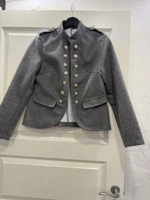 Grå rock med dekorativa knappar - Snygg grå kavaj med silverfärgade knappar i två rader framtill. Modellen är figurnära med lång ärm. 