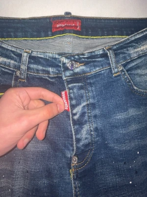 Blå Dsquared2 ICON jeans med slitningar - Säljer ett par blå Dsquared2 ICON jeans med coola slitningar och färgstänk på benen. Klassisk femficksmodell med Dsquared2-tag på fickan och stor ICON-label bak i midjan. Jeansen har en rak passform och är gjorda i jeansmaterial med snygga detaljer.