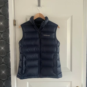 Mörkblå dunväst Peak Performance S - Snygg mörkblå dunväst från Peak Performance i storlek S. Västen har hög krage, diskret logga på bröstet och dragkedja framtill. Perfekt för lager-på-lager och chill höst- eller vinterstil.