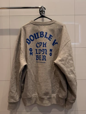 Grå sweatshirt från Wood Wood - Säljer en grå sweatshirt från Wood Wood med blått tryck på ryggen och ett W på bröstet. Tröjan har en loose passform, rund hals och är tillverkad i mjuk bomull. Stora i storleken, jag är 180 och den passsar. Nypris ca 1000kr säljes för 300kr