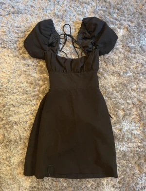Svart kort klänning från Zara - Supersnygg svart klänning från Zara med puffärmar och snörning i ryggen. Modellen är kort och har en figurnära passform med fyrkantig urringning. Materialet känns som bomull och polyester, vilket gör den bekväm och lätt att bära. Perfekt för dig som gillar trendiga detaljer.