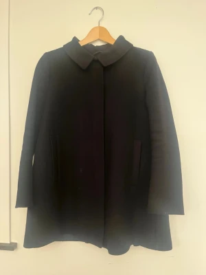 Svart ullkappa från COS - Stilren svart kappa från COS i ullmix med klassisk krage och dolda fickor. Jackan har diskret dragkedja och är helfodrad. 