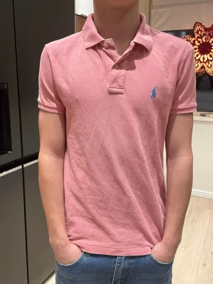 Rosa pikétröja från Ralph Lauren - Säljer en klassisk rosa pikétröja från Ralph Lauren med blå broderad logga på bröstet. Tröjan har korta ärmar, krage och två knappar vid halsen. Perfekt för dig som gillar stilrena och tidlösa plagg.
