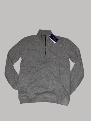Grå half zip tröja från Polo Ralph Lauren - Snygg grå långärmad tröja från Polo Ralph Lauren med half zip och klassisk krage. Tröjan har ett broderat logomärke på bröstet och är tillverkad i mjuk bomull. Perfekt för dig som gillar stilrena och bekväma plagg med premiumkänsla.