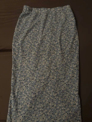 Blommig blå långkjol med slits - Säljer en blå långkjol med vitt blommigt mönster och diskreta svarta detaljer. Kjolen har en rak form och en slits baktill för extra rörelsefrihet. Perfekt för dig som gillar en feminin och chill stil. Materialet känns mjukt och lätt mot huden.