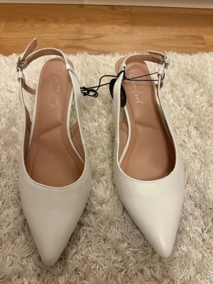 Vita pumps med spetsig tå från New Look - Snygga vita pumps från New Look med spetsig tå och smal klack. Skorna har en öppen häl med justerbart spänne och är gjorda i slätt syntetmaterial. Perfekt för dig som gillar stilrena och eleganta skor med modern touch. Helt nya 