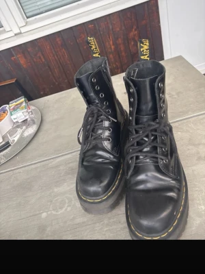 Svarta Dr. Martens boots med plattform - Säljer ett par svarta Dr. Martens boots med klassisk gul söm runt sulan och grov plattform. Skorna har snörning, rund tå och är tillverkade i blankt skinn. Ikonisk AirWair-flik baktill. Perfekt för dig som vill ha en edgy stil.