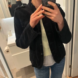 Svart manchesterjacka med knappar - Napoleonjacka / 2000s. Säljer en svart manchesterjacka med coola detaljer som axelklaffar och stora knappar framtill. Jackan har ståkrage, fickor med lock på bröstet och är figursydd för en snygg passform. Perfekt att slänga på sig över en hoodie eller t-shirt. Strl 38