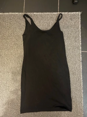 Svart ärmlös kortklänning - En enkel svart klänning med smala axelband och ärmlös design. Klänningen är kort och har en rak, tight passform som framhäver siluetten. Perfekt för dig som gillar minimalistisk stil och vill ha en klassisk bas i garderoben.