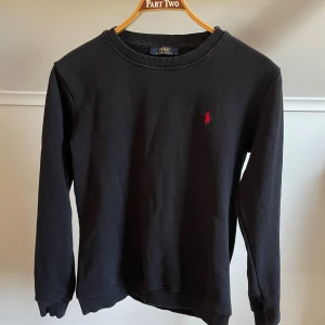 Svart sweatshirt från Polo Ralph Lauren - Klassisk svart sweatshirt från Polo Ralph Lauren med rund halsringning och röd broderad logga på bröstet. Perfekt för en stilren och avslappnad look.