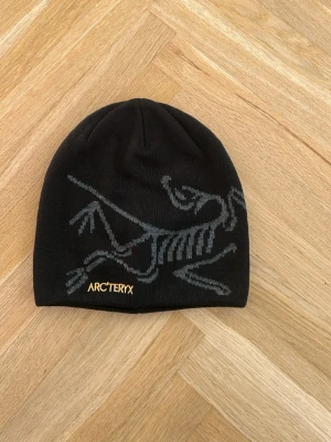 Svart mössa från Arc'teryx - Svart mössa från Arc'teryx med grått skelettmönster på framsidan och gul logotyp. Snygg och enkel design som passar perfekt för kalla dagar. Materialet är stickat och känns mjukt mot huvudet.