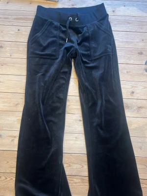 Svarta velourbyxor Juicy Couture XS - Svarta velourbyxor från Juicy Couture i storlek XS. Byxorna har bred midja med snörning, stora fickor fram och klassisk Juicy-text med paljetter på baksidan. De är uppsydda vilket gör de på gränsen till för korta för mig som är ca 167 men det borde gå enkelt att sprätta. Säljer för att de inte kommer till användning. Buda gärna! 