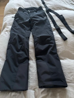 Svarta skidbyxor Peak Performance XS - Svarta täckbyxor från Peak Performance i storlek XS. Byxorna har raka ben, förstärkta partier vid bensluten och medföljande svarta hängslen. De är tillverkade i slitstarkt syntetmaterial och har dubbla knappar i midjan samt dragkedja.