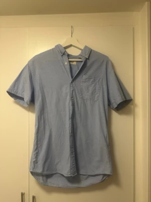 Ljusblå kortärmad skjorta från Produkt - Fräsch ljusblå skjorta från Produkt i storlek M. Skjortan har korta ärmar, klassisk krage med knappar och en bröstficka. Nedersta knappen är ditsydd med svart tråd.