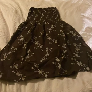 Brun blommig axelbandslös klänning - Säljer en brun axelbandslös klänning med vitt blommigt broderat mönster. Klänningen har en smockad överdel och ett utställt kort kjolparti. Materialet känns lätt och luftigt, perfekt för varmare dagar. Söt och romantisk stil med fina detaljer.Funksr att använda som kjol med eftersom det blir en ganska kort klänning,sprund i båda sidorna.Köpt i Thailand.