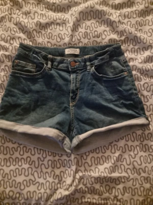Blå denimshorts från Lindex - Säljer ett par klassiska blå denimshorts från Lindex med uppvikta benslut och femficksmodell. Shortsen har normal passform och är tillverkade i bomull. Perfekta för varma dagar och enkla att matcha med allt.