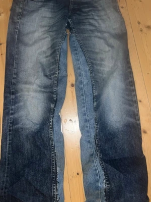 Blå raka jeans med slitna detaljer - Säljer ett par blå jeans med raka ben och slitna detaljer längs sidorna och vid bensluten. Jeansen har klassisk femficksdesign och är tillverkade i robust denim. Färgen är mörkblå med ljusare tvättade partier för en cool, använd look.