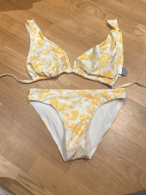 Blommig bikini från Kappahl - Säljer en fräsch bikini från Kappahl med vit botten och gula blommor. Överdelen har knyt i nacken och underdelen är klassisk med blommigt mönster. Materialet är mjukt och syntetiskt, perfekt för strandhäng. Supersnygg färgkombo och somrig vibe! överdel L underdel XS