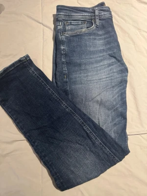 Jack & Jones Regular Clark jeans blå - Säljer ett par Jack & Jones Regular Clark jeans i klassisk blå tvätt. Jeansen är i okej skick där jag använt dom hyffsat mycket. Modellen har rak passform, fem fickor och snygga slitningar framtill. Jeansen är tillverkade i bomull och har normal midja. Perfekt för dig som gillar en enkel och stilren look.
