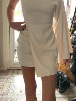 Vit Adoore one shoulder kortklänning i satin - Snygg vit kortklänning i satin från Adoore med one shoulder-design och en lång, vid ärm. Klänningen har en omlottliknande detalj framtill och sitter tight över kroppen. Använd 1 gång storlek xs.