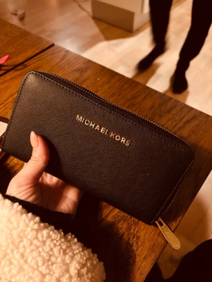 Svart plånbok från Michael Kors - Säljer en stilren svart plånbok/väska från Michael Kors med guldfärgad dragkedja och logga. Plånboken är rektangulär och har flera fack inuti. Aldrig använd.