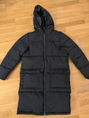 Svart lång pufferjacka från Object - Lång svart pufferjacka från Object med huva och stora fickor framtill. Jackan har dragkedja hela vägen och en clean, quiltad look. Perfekt för kalla dagar när du vill hålla dig varm och stilsäker.