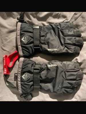 Svarta vinterhandskar från Hestra i vuxenstorlek 7 - Svarta vinterhandskar från Hestra med justerbart band runt handleden och röd handledsrem. Handskarna har förstärkta partier i handflatan och är fodrade för extra värme. Perfekta för kalla dagar och vinteraktiviteter. Sparsamt använda!